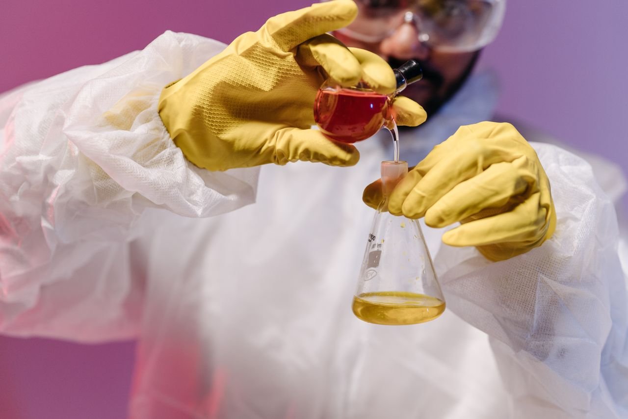 Scientist in protective gear pouring colored liquid into flask in a laboratory setting.